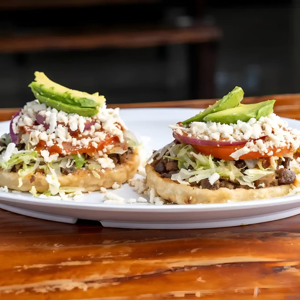 Two Mexican sopes topped with meat, lettuce, cheese, tomato, onion, and avocado slices.