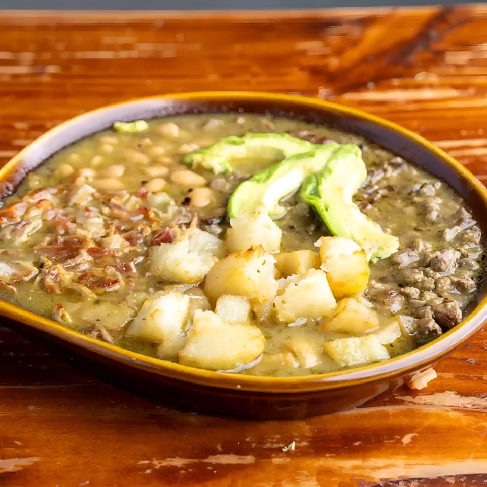 A bowl of Mexican pozole with white beans, potatoes, meat, and avocado slices.